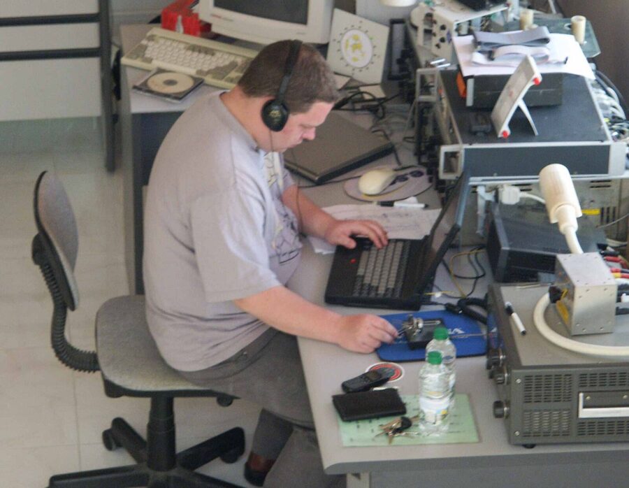 Amateur radio operator wearing headphones at a desk station with transceivers, a laptop log, and a Morse paddle
